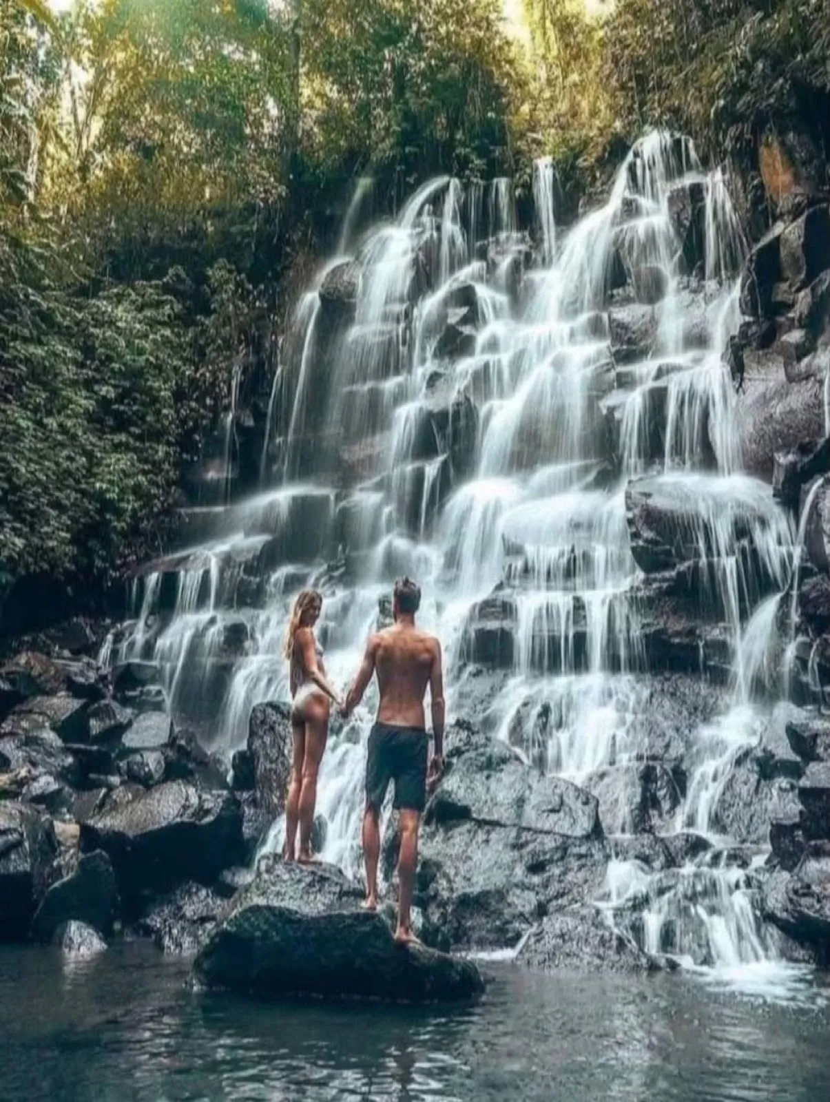 A couple on a rock in the pool, facing the wide curtain of Kanto Lampo Waterfall