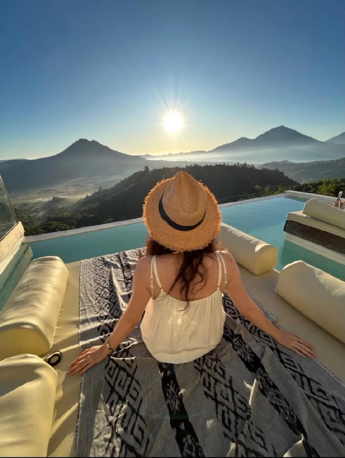 A guest in a straw hat on a daybed beside an infinity pool, facing a volcano and a lake