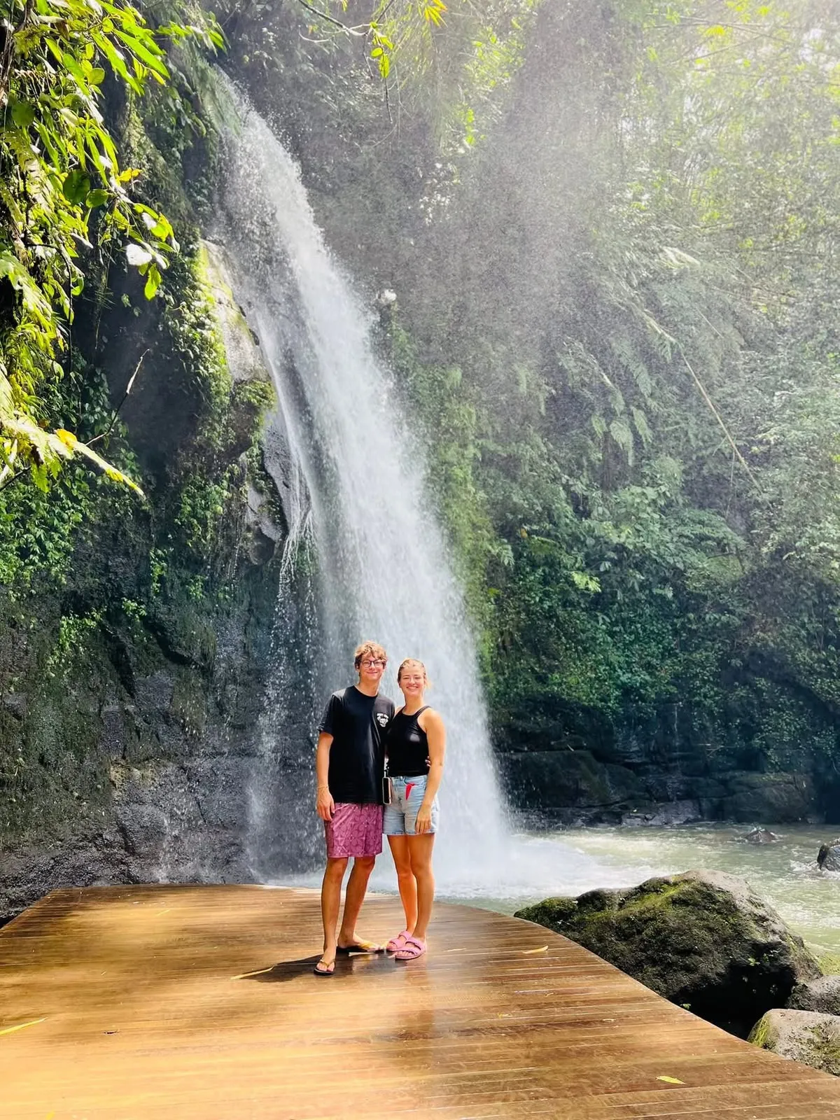 A couple on a wooden platform in front of a tall forest waterfall at Ulu Petanu