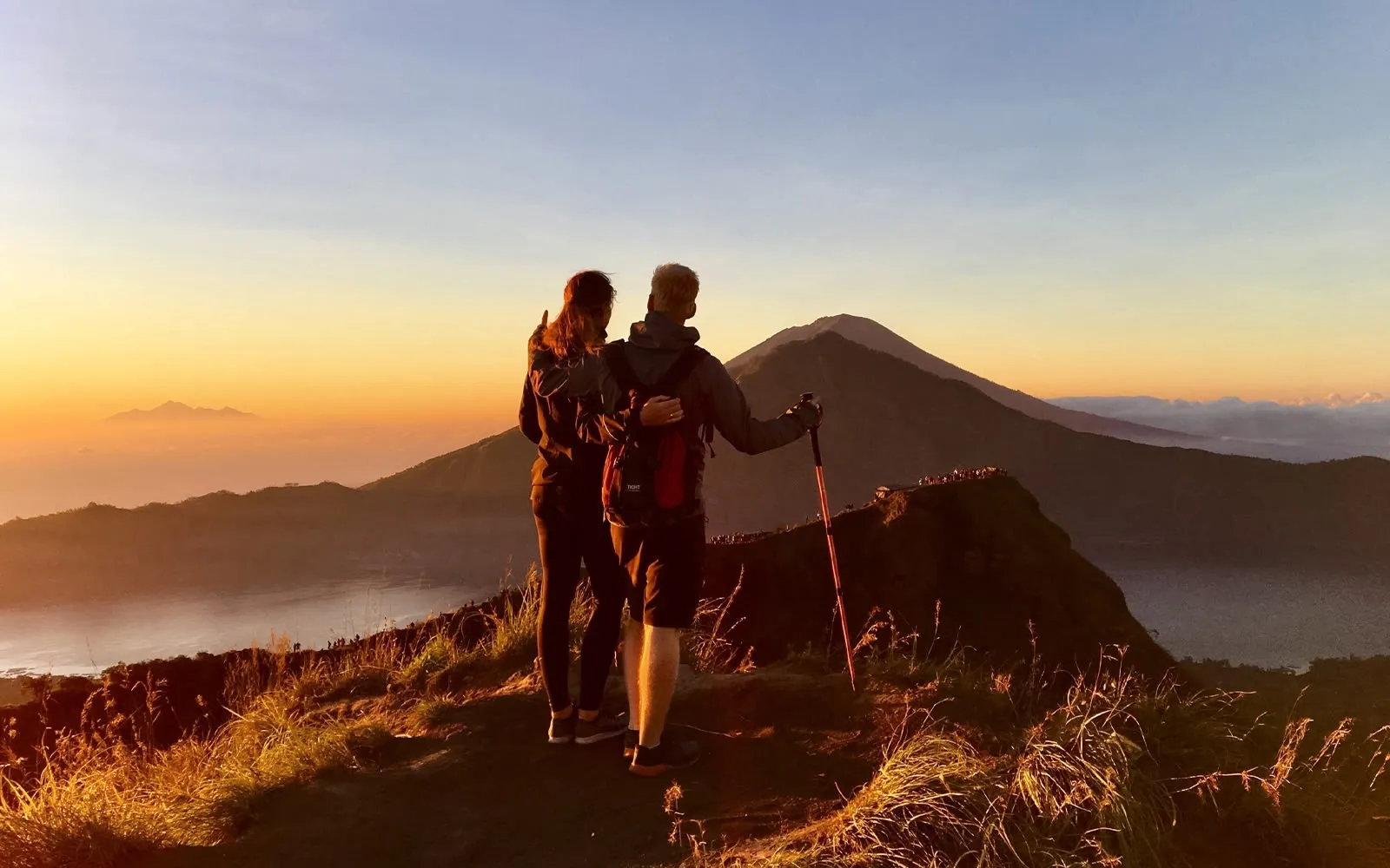 Two hikers with a trekking pole on a ridge at sunrise, a lake and a volcano behind them