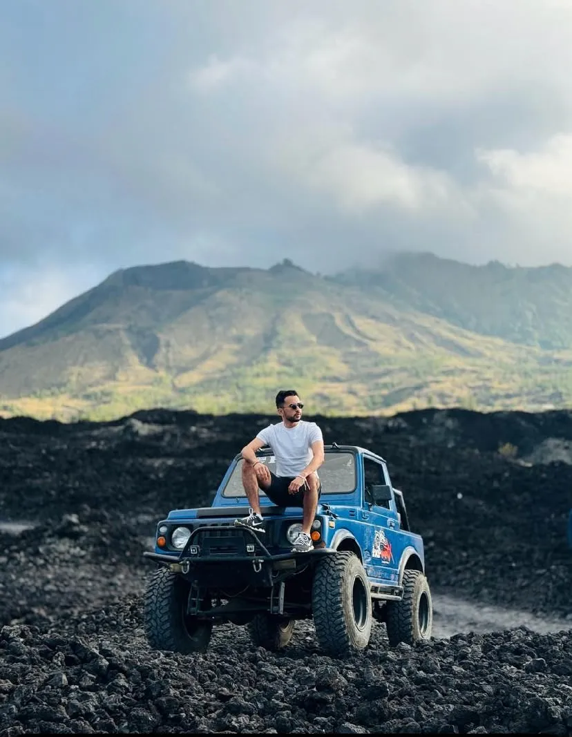 Guest seated on a blue jeep on black volcanic rock, with Mount Batur behind