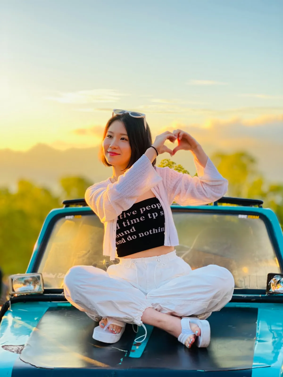 Guest making a heart shape while seated on a turquoise jeep at sunset
