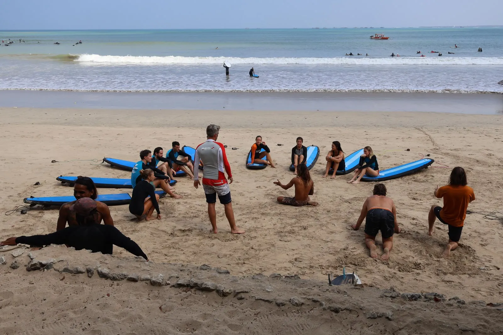 Surf instructor standing with a class seated on the sand beside blue boards
