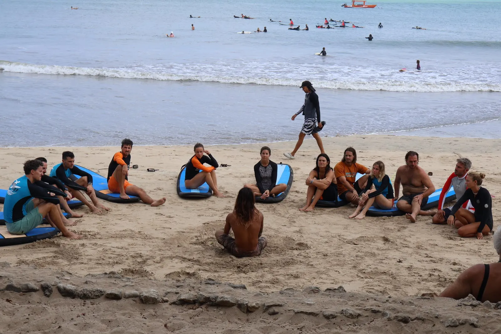 A group surf class sitting in a circle on the beach with blue boards